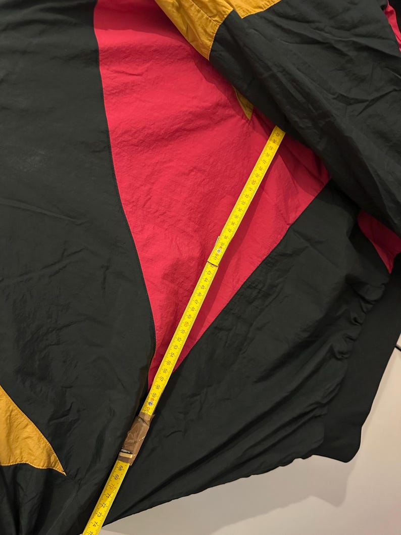 K&ouml;nnte beinhalten: Eine Vintage-Windjacke in Schwarz, Rot und Gold. Die Jacke wird mit einem gelben Ma&szlig;band gemessen. Die Windjacke hat ein Farbblock-Design mit einem geometrischen Muster. Die Jacke ist aus leichtem Material gefertigt.