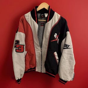 Puede incluir: Una chaqueta bomber vintage Nike Air Jordan con un esquema de color rojo, blanco y negro. La chaqueta presenta un gran logotipo "AIR" con una silueta de jugador de baloncesto en la espalda, un "23" en una manga y un swoosh de Nike en la otra manga.