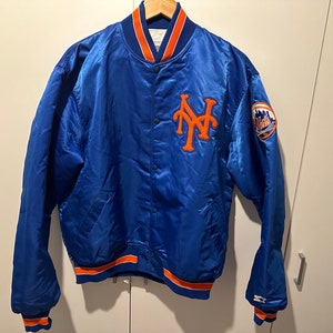 Può includere: Una giacca da baseball in satin blu con bordini arancioni e bianchi. La giacca presenta un grande logo "NY" ricamato sul petto sinistro e una toppa più piccola con una palla da baseball e le parole "New York" sulla manica destra.