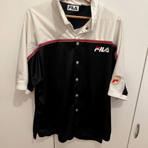 Peut inclure: Une chemise boutonn&eacute;e avec une partie sup&eacute;rieure blanche et une partie inf&eacute;rieure noire. La chemise a des manches courtes et un d&eacute;tail de rayure rouge. Le logo Fila est visible sur la poitrine et le col. La chemise est suspendue &agrave; un cintre en bois.