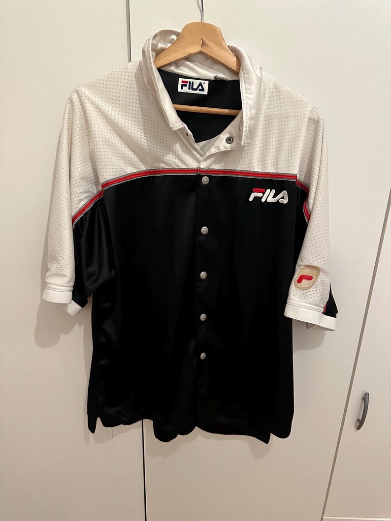 Peut inclure: Une chemise boutonn&eacute;e avec une partie sup&eacute;rieure blanche et une partie inf&eacute;rieure noire. La chemise a des manches courtes et un d&eacute;tail de rayure rouge. Le logo Fila est visible sur la poitrine et le col. La chemise est suspendue &agrave; un cintre en bois.