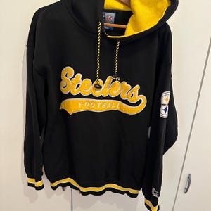 Könnte beinhalten: Schwarzer Kapuzenpullover mit gelb gefütterter Kapuze und dem Wort "Steelers" in gelber Schrift auf der Vorderseite. Das Wort "FOOTBALL" steht darunter. Die Bündchen und der untere Saum haben gelbe Streifen. Ein Aufnäher mit Logo befindet sich am Ärmel.
