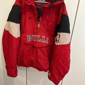 Puede incluir: Una chaqueta tipo cortavientos de la marca Starter en colores rojo, negro y blanco, con capucha, cierre de media cremallera y la palabra BULLS bordada en el bolsillo central. Incluye un parche con el logo de la NBA en la manga y forro acolchado.