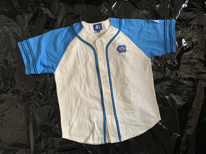 May include: White baseball jersey with blue sleeves and a black and blue striped trim. The jersey has a button-up front and a small embroidered logo on the left chest.