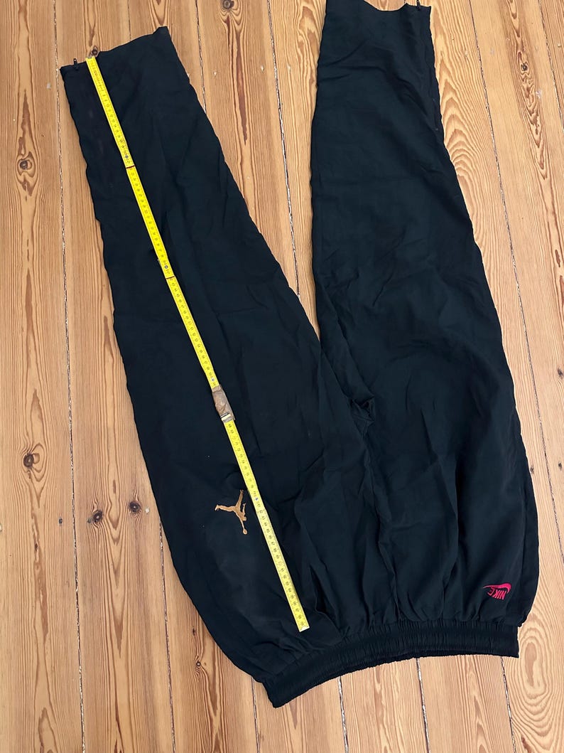K&ouml;nnte beinhalten: Schwarze Sporthose mit goldenem Jordan-Logo und rotem Nike-Logo. Ein gelbes Ma&szlig;band liegt &uuml;ber der Hose. Die Hose besteht aus leichtem Material und hat einen elastischen Bund.
