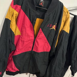 K&ouml;nnte beinhalten: Schwarze, rote und goldfarbene Jordan-Jacke mit durchgehendem Rei&szlig;verschluss. Die Jacke hat ein Colorblock-Design mit dem Jordan-Logo auf der Brust und am &Auml;rmel. Die Jacke h&auml;ngt an einem schwarzen Kleiderb&uuml;gel.
