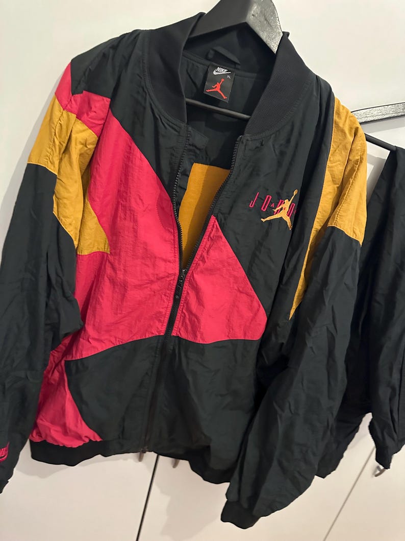 K&ouml;nnte beinhalten: Schwarze, rote und goldfarbene Jordan-Jacke mit durchgehendem Rei&szlig;verschluss. Die Jacke hat ein Colorblock-Design mit dem Jordan-Logo auf der Brust und am &Auml;rmel. Die Jacke h&auml;ngt an einem schwarzen Kleiderb&uuml;gel.