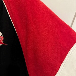 May include: Close-up of a red and black jacket sleeve. The sleeve is red with a white stripe and a black section. The jacket appears to be made of a soft, textured material.
