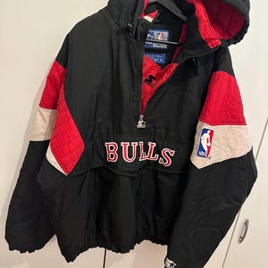 Op de afbeelding: Zwarte, rode en witte Starter jas met capuchon. De jas heeft het woord "BULLS" over de borst en een NBA-logo. De jas heeft een halve ritssluiting en een Starter-logo op de mouw.