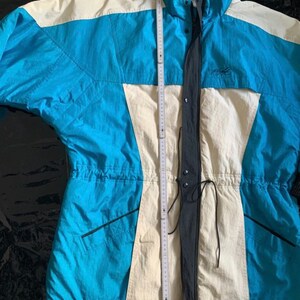 May include: A blue and white ski jacket with a black zipper and drawstring waist. The jacket has a high collar and a logo on the chest.