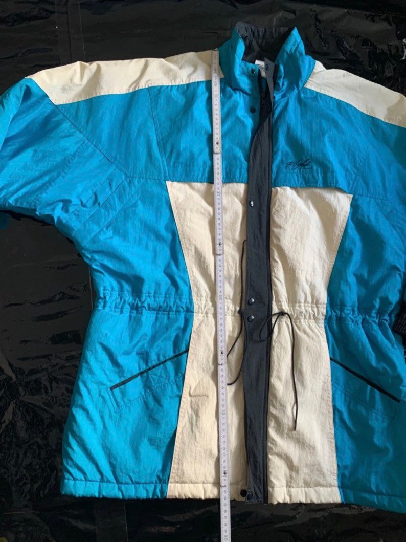 May include: A blue and white ski jacket with a black zipper and drawstring waist. The jacket has a high collar and a logo on the chest.
