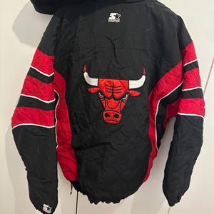 Op de afbeelding: Zwarte en rode Chicago Bulls Starter jas met capuchon. De jas heeft het Bulls-logo op de rug en rode strepen op de mouwen. Het Starter-logo is zichtbaar op de borst en de mouw.