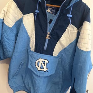 May include: A vintage light blue, navy, and white Starter pullover jacket with a hood. The jacket features a large pocket with the white and navy "NC" logo. The jacket has a zipper and drawstrings. The jacket is made of a lightweight material.