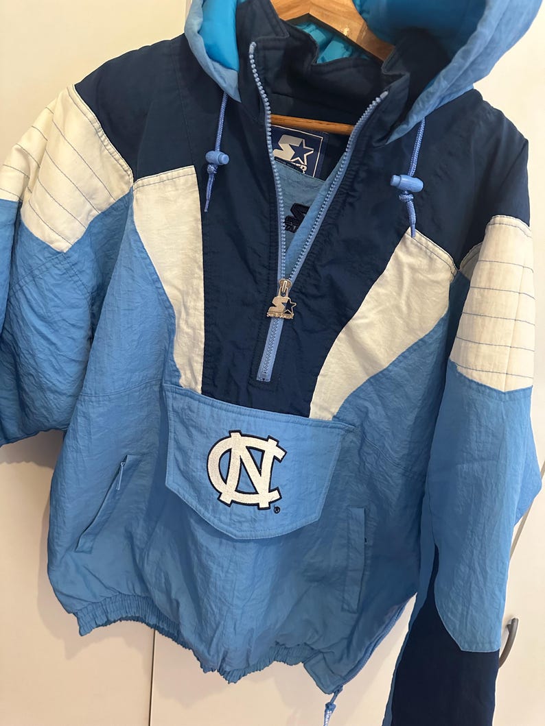 May include: A vintage light blue, navy, and white Starter pullover jacket with a hood. The jacket features a large pocket with the white and navy "NC" logo. The jacket has a zipper and drawstrings. The jacket is made of a lightweight material.