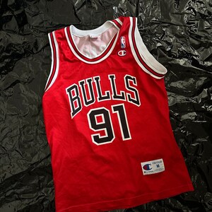 May include: Red and black basketball jersey with the word "BULLS" and the number "91" on the front. The jersey is a size medium and has a Champion logo on the front.