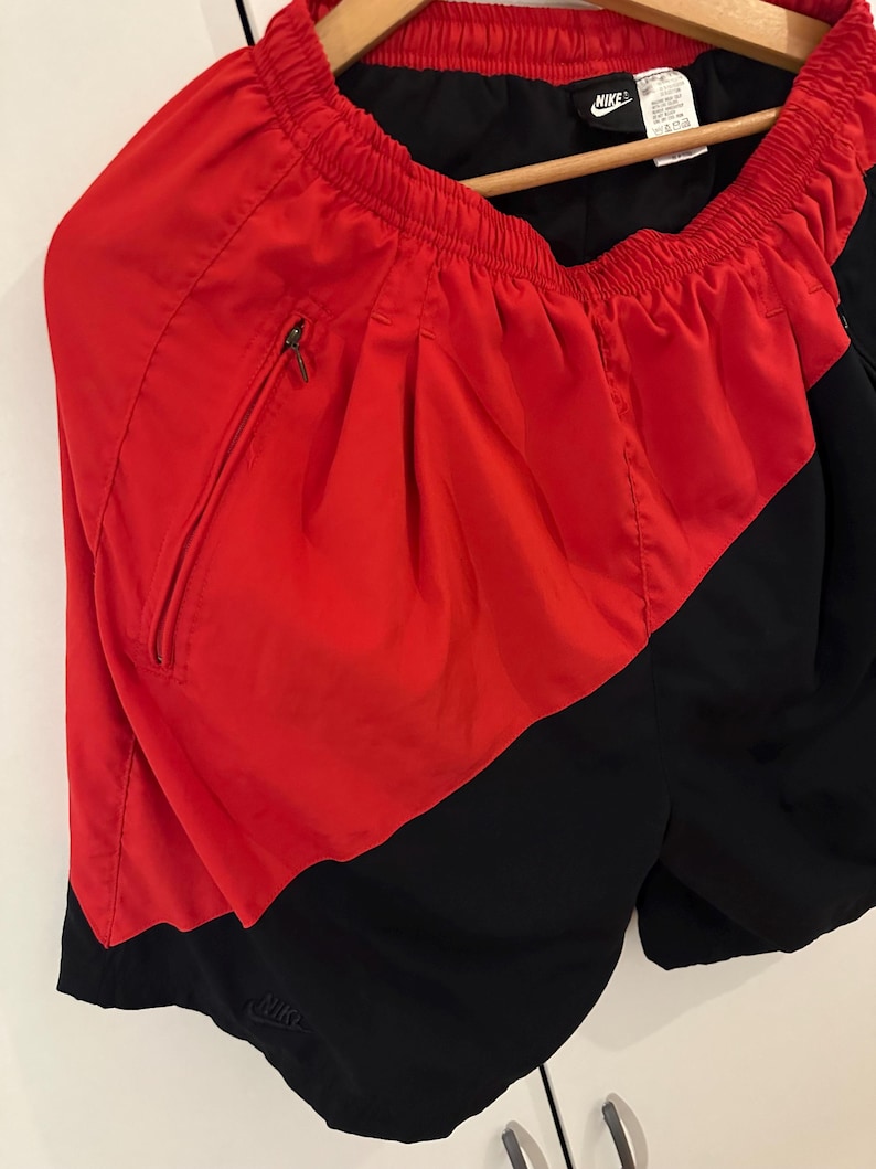 May include: Red and black athletic shorts with a Nike logo on the side and a zippered pocket. The shorts have an elastic waistband.
