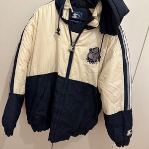 Puede incluir: Chaqueta Starter crema y azul marino con capucha y cremallera completa. La chaqueta presenta un parche con el logotipo de un bulldog y el logotipo de Starter en la manga. Diseño a rayas en las mangas.