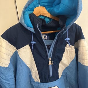 May include: A blue, white, and navy blue Starter pullover jacket with a hood. The jacket features a zipper closure, a front pocket, and the University of North Carolina logo. The jacket is hanging on a wooden hanger.