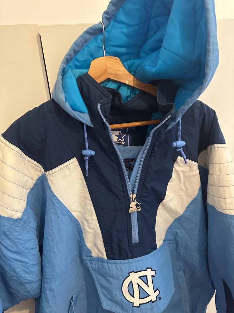 May include: A blue, white, and navy blue Starter pullover jacket with a hood. The jacket features a zipper closure, a front pocket, and the University of North Carolina logo. The jacket is hanging on a wooden hanger.
