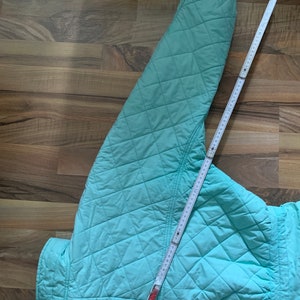 May include: A light blue quilted jacket with a diamond pattern. The jacket is laid flat on a wooden surface with a measuring tape.