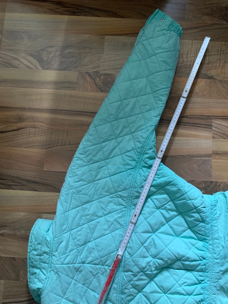 May include: A light blue quilted jacket with a diamond pattern. The jacket is laid flat on a wooden surface with a measuring tape.