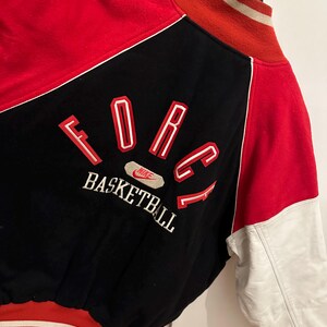 May include: A vintage black, red, and white Nike Force Basketball jacket. The jacket features a black body with red accents on the shoulders and sleeves. The word "FORCE" is embroidered in red with white outlines on the back, along with "BASKETBALL". The sleeves are white.
