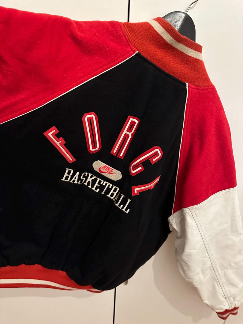 May include: A vintage black, red, and white Nike Force Basketball jacket. The jacket features a black body with red accents on the shoulders and sleeves. The word "FORCE" is embroidered in red with white outlines on the back, along with "BASKETBALL". The sleeves are white.