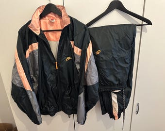 May include: Black and grey Nike tracksuit with peach and gold detailing. The jacket features a full zip and the Nike logo in gold. The trousers are black with a white and peach stripe. The tracksuit is hanging on a black hanger.
