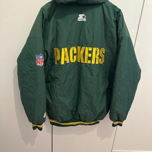 Könnte beinhalten: Grün-gelbe Packers-Jacke mit Kapuze. Auf der Rückseite steht das Wort "PACKERS" in großen gelben Buchstaben. Die Jacke hat außerdem das NFL-Logo und das Starter-Logo. Sie hat einen gelb-grünen Streifen.