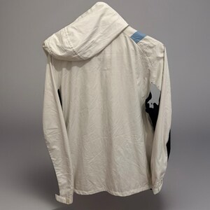 Puede incluir: Chaqueta con capucha color crema con un acento azul claro en el hombro y detalles negros en la manga. La chaqueta est&aacute; hecha de un material ligero y tiene una cremallera completa.