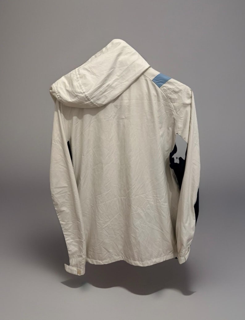 Puede incluir: Chaqueta con capucha color crema con un acento azul claro en el hombro y detalles negros en la manga. La chaqueta est&aacute; hecha de un material ligero y tiene una cremallera completa.