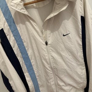 Puede incluir: Chaqueta cortavientos Nike blanca con capucha y cierre de cremallera. La chaqueta presenta un dise&ntilde;o de rayas azul claro y azul marino en la parte delantera y detalles en azul marino en los laterales. Un logotipo negro de Nike en el pecho.