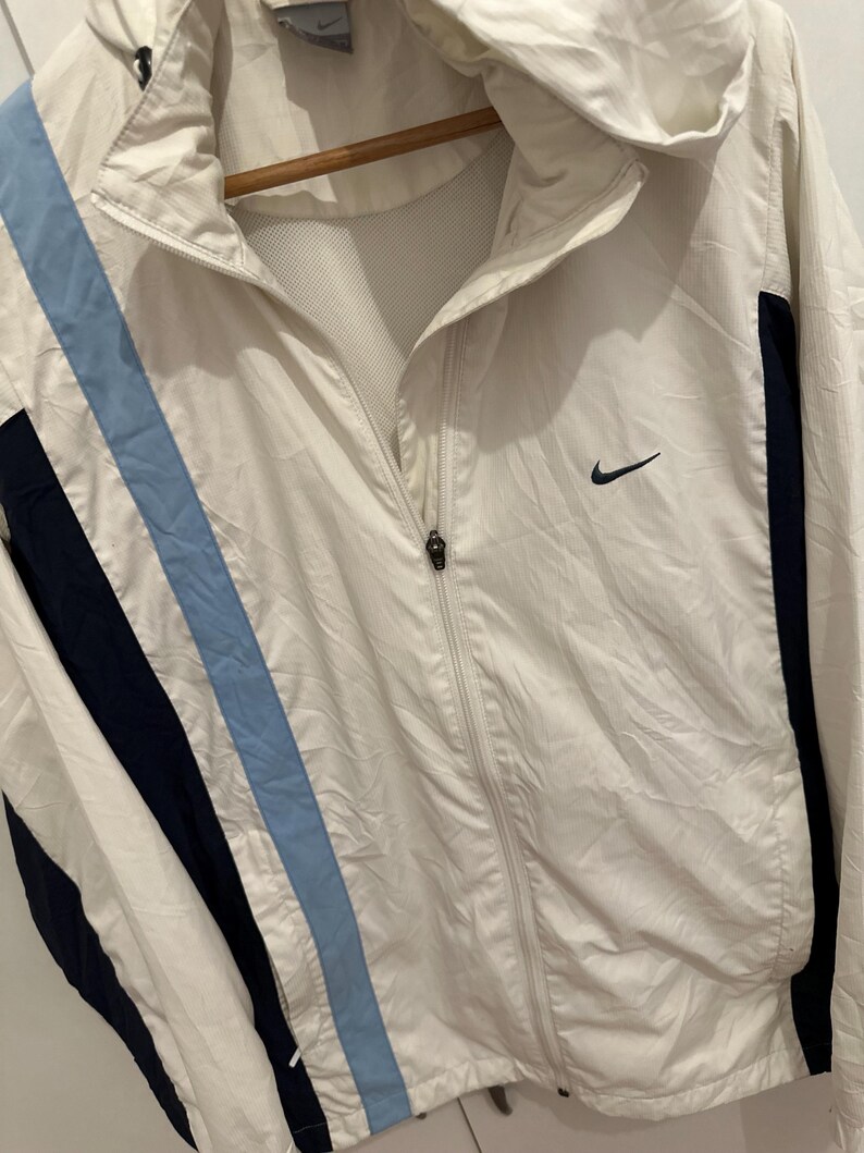 Puede incluir: Chaqueta cortavientos Nike blanca con capucha y cierre de cremallera. La chaqueta presenta un dise&ntilde;o de rayas azul claro y azul marino en la parte delantera y detalles en azul marino en los laterales. Un logotipo negro de Nike en el pecho.