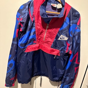 Puede incluir: Una chaqueta cortavientos azul, roja y blanca con un patrón geométrico y la palabra "NIKE" en la parte delantera.