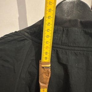 K&ouml;nnte beinhalten: Eine schwarze Jacke wird mit einem gelben Ma&szlig;band gemessen. Das Ma&szlig;band zeigt eine L&auml;nge von etwa 200 cm. Die Jacke h&auml;ngt an einem schwarzen Kleiderb&uuml;gel.