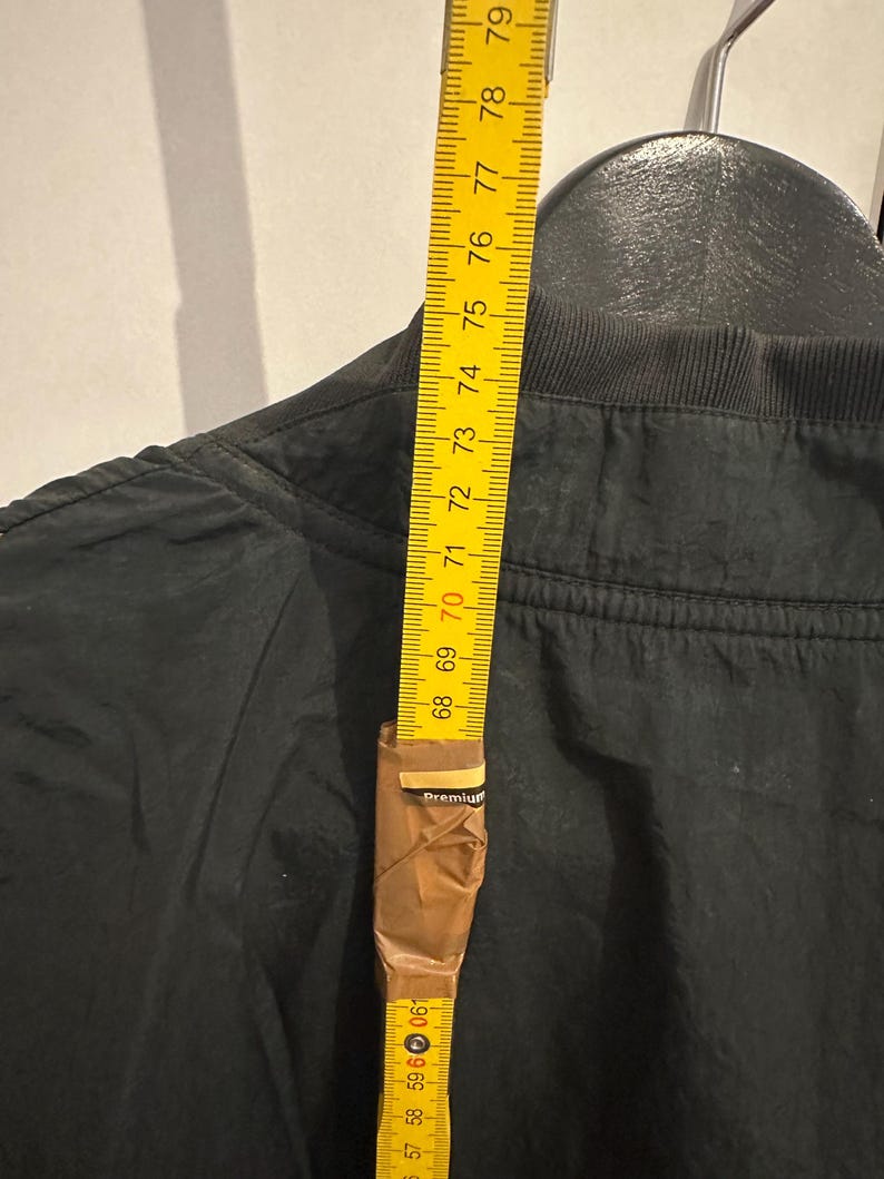 K&ouml;nnte beinhalten: Eine schwarze Jacke wird mit einem gelben Ma&szlig;band gemessen. Das Ma&szlig;band zeigt eine L&auml;nge von etwa 200 cm. Die Jacke h&auml;ngt an einem schwarzen Kleiderb&uuml;gel.