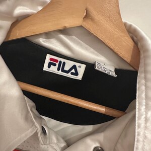 Peut inclure: Veste Fila blanche et noire avec une bande rouge, suspendue &agrave; un cintre en bois. La veste a une fermeture boutonn&eacute;e et le logo Fila est visible. La veste est en polyester.