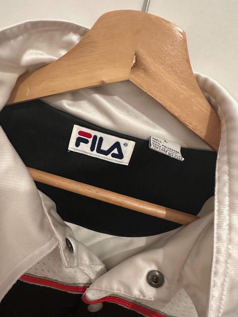 Peut inclure: Veste Fila blanche et noire avec une bande rouge, suspendue &agrave; un cintre en bois. La veste a une fermeture boutonn&eacute;e et le logo Fila est visible. La veste est en polyester.