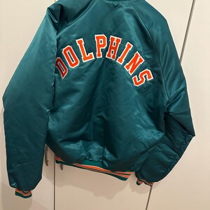 Op de afbeelding: Een teal satijnen bomberjack met het woord DOLPHINS in oranje borduursel met een witte omlijning op de achterkant. Het jack heeft een geribbelde kraag, boorden en zoom met oranje en witte strepen en hangt aan een houten kledinghanger.
