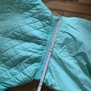 May include: A light blue quilted jacket with a zipper closure. The jacket is laid flat on a wooden surface with a measuring tape extending from the bottom of the jacket.