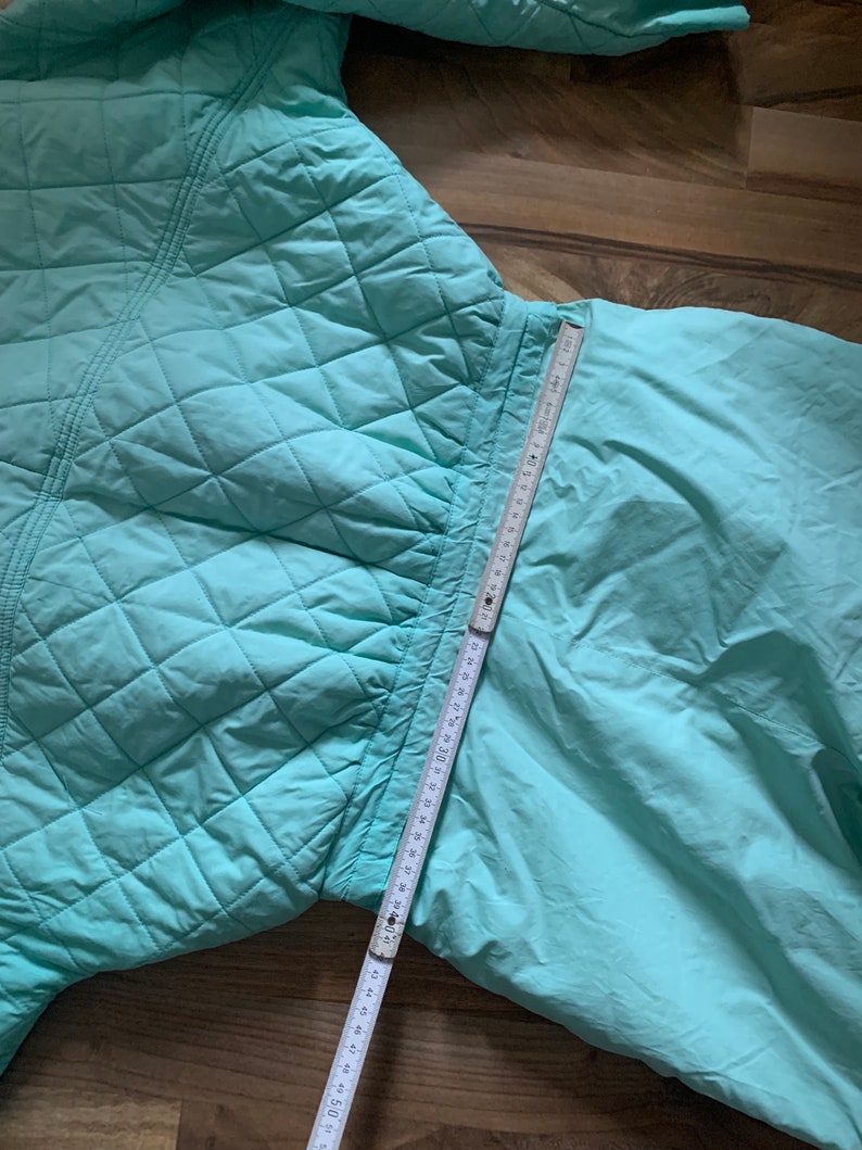 May include: A light blue quilted jacket with a zipper closure. The jacket is laid flat on a wooden surface with a measuring tape extending from the bottom of the jacket.