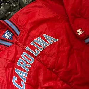 May include: A red Starter brand satin bomber jacket with a blue and white striped collar and cuffs. The jacket features a large blue embroidered "CAROLINA" on the front.