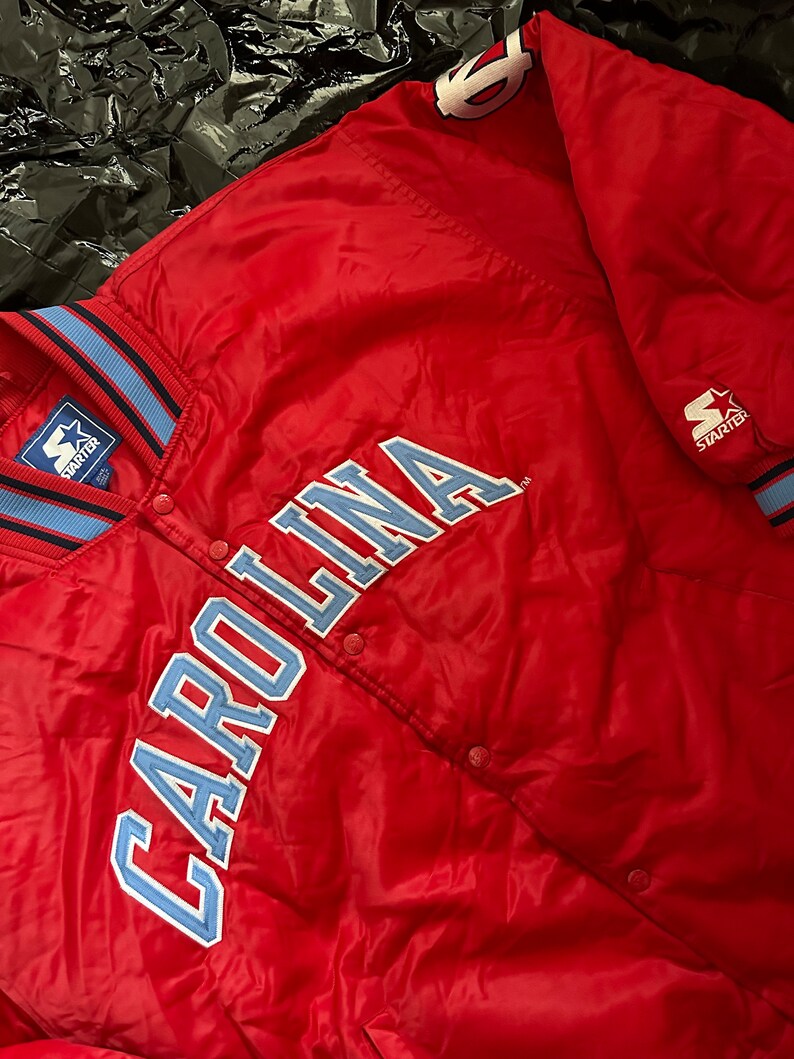 May include: A red Starter brand satin bomber jacket with a blue and white striped collar and cuffs. The jacket features a large blue embroidered "CAROLINA" on the front.