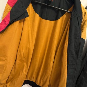 K&ouml;nnte beinhalten: Eine schwarze, goldene und rosa Bomberjacke mit durchgehendem Rei&szlig;verschluss. Die Jacke hat ein goldenes Innenfutter und einen schwarzen Kragen und B&uuml;ndchen. Das Nike-Logo ist oben auf dem R&uuml;cken sichtbar.