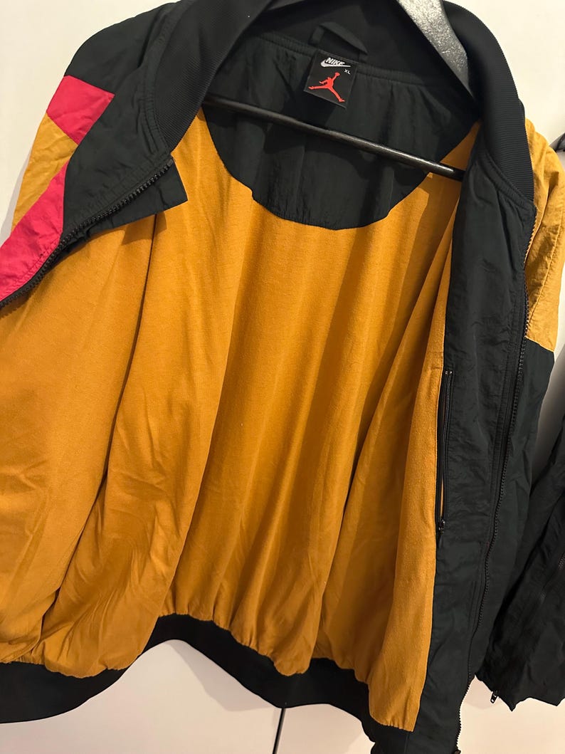K&ouml;nnte beinhalten: Eine schwarze, goldene und rosa Bomberjacke mit durchgehendem Rei&szlig;verschluss. Die Jacke hat ein goldenes Innenfutter und einen schwarzen Kragen und B&uuml;ndchen. Das Nike-Logo ist oben auf dem R&uuml;cken sichtbar.