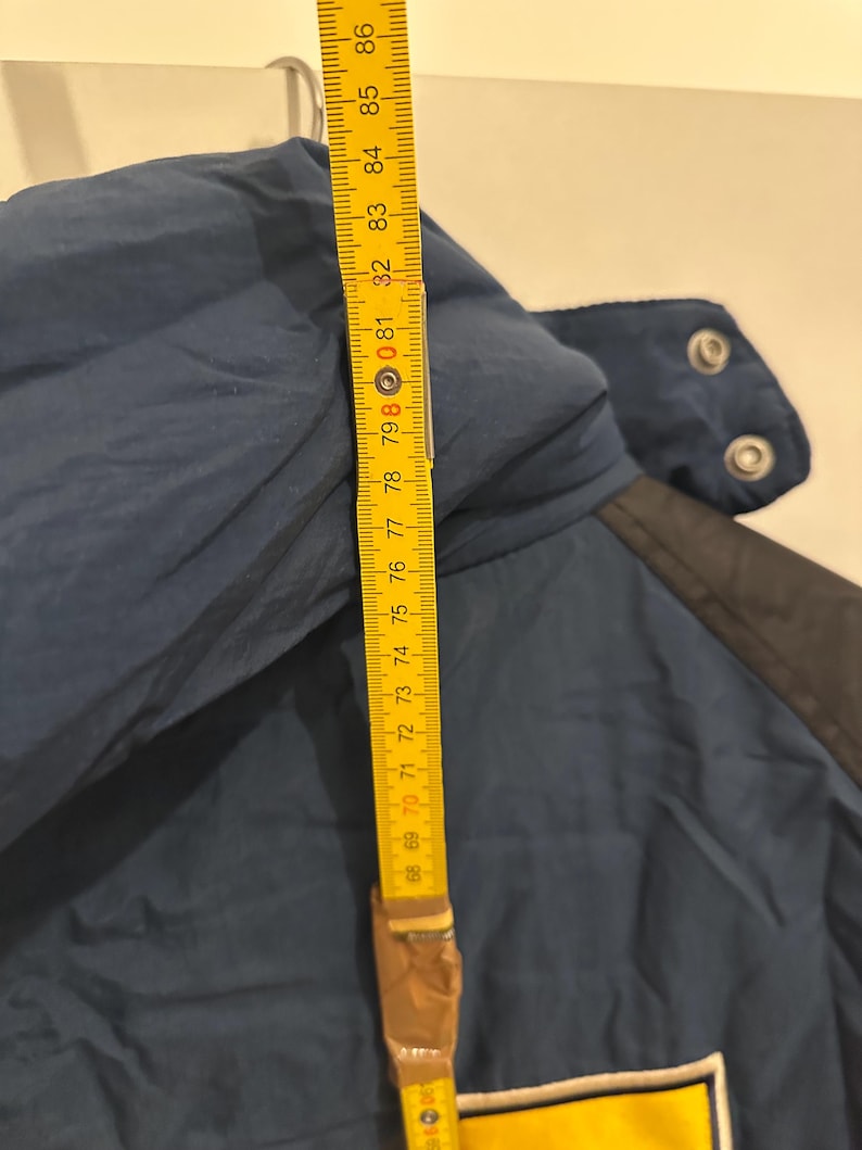 May include: A navy blue jacket with a yellow and white patch. A yellow measuring tape shows a length of approximately 30 inches. The jacket has a black collar and snap closures.