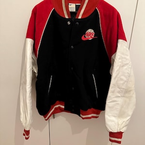 May include: Black, red, and white varsity jacket with a Nike logo. The jacket has a black body, red sleeves, and white sleeves. The collar and cuffs are red and white striped. The Nike logo is embroidered on the chest.