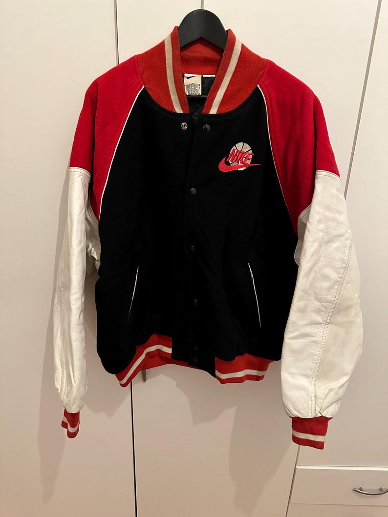 May include: Black, red, and white varsity jacket with a Nike logo. The jacket has a black body, red sleeves, and white sleeves. The collar and cuffs are red and white striped. The Nike logo is embroidered on the chest.