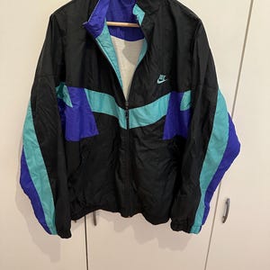 May include: A black, teal, and purple windbreaker jacket featuring a full-length front zipper, a high collar, and long sleeves with elastic cuffs. The Nike logo is embroidered in teal on the upper left chest area of the garment.