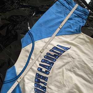 May include: A white and light blue baseball jersey with the text "NORTH CAROLINA" embroidered in blue on the front. A measuring tape is visible in the image.